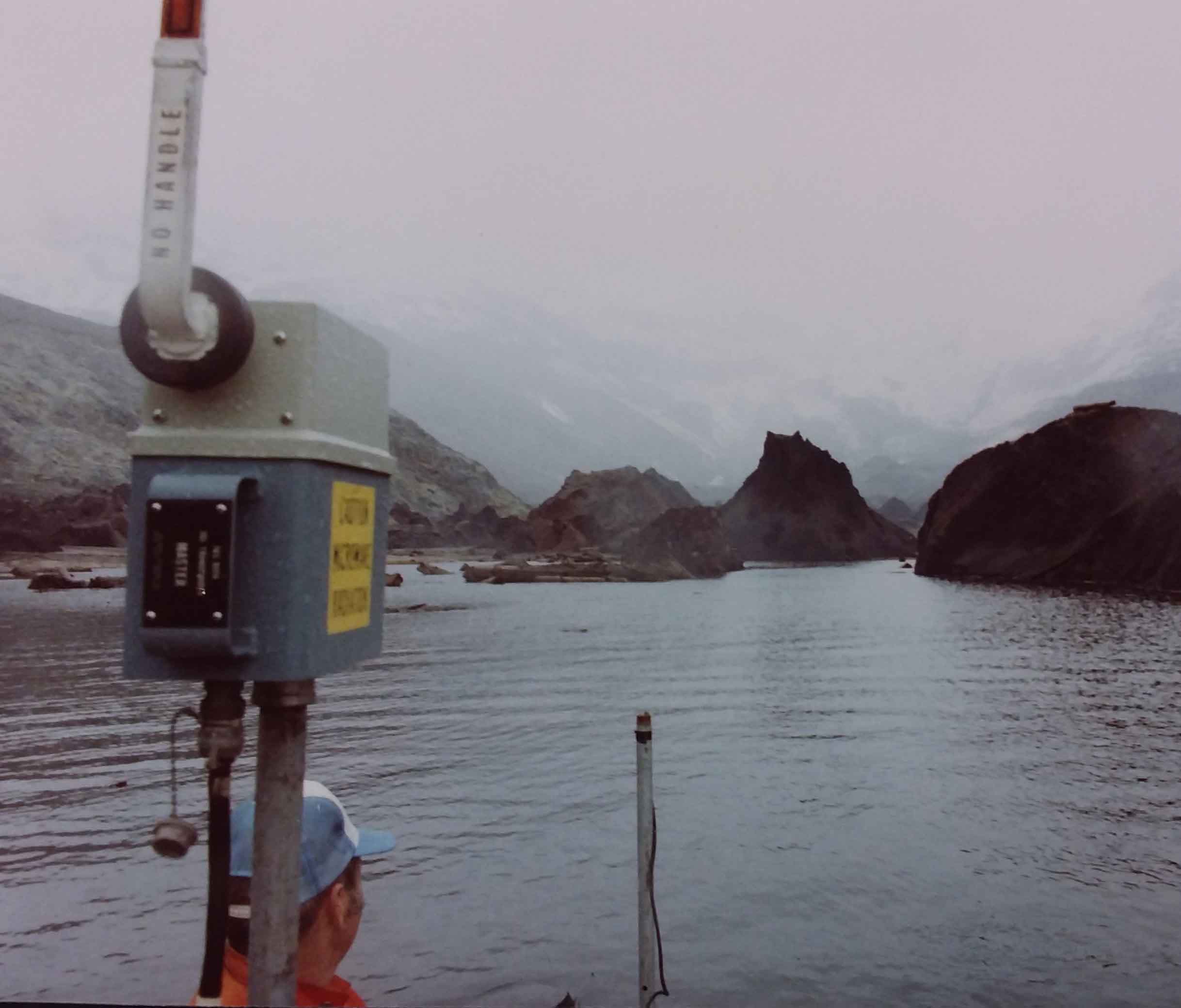 May 5, 1981 - Leonard Nelson in aluminum boat in Bear Cove, West Arm of Spirit Lake with debris avalanche hummocks. Microwave trisponder range finder in boat for use in bathymetric mapping. USGS K. Tanner